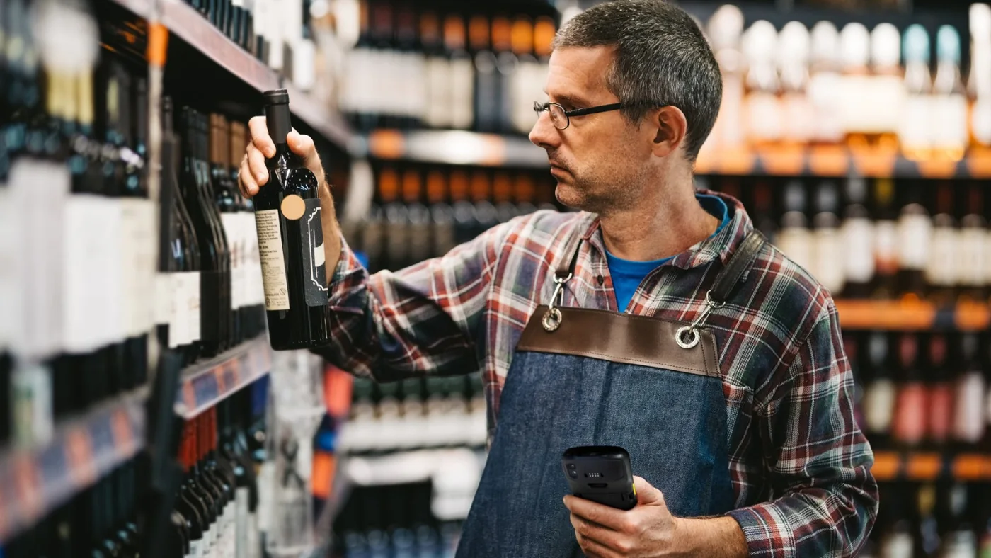 Dipendente del negozio che controlla una bottiglia di vino sullo scaffale con un computer mobile Zebra